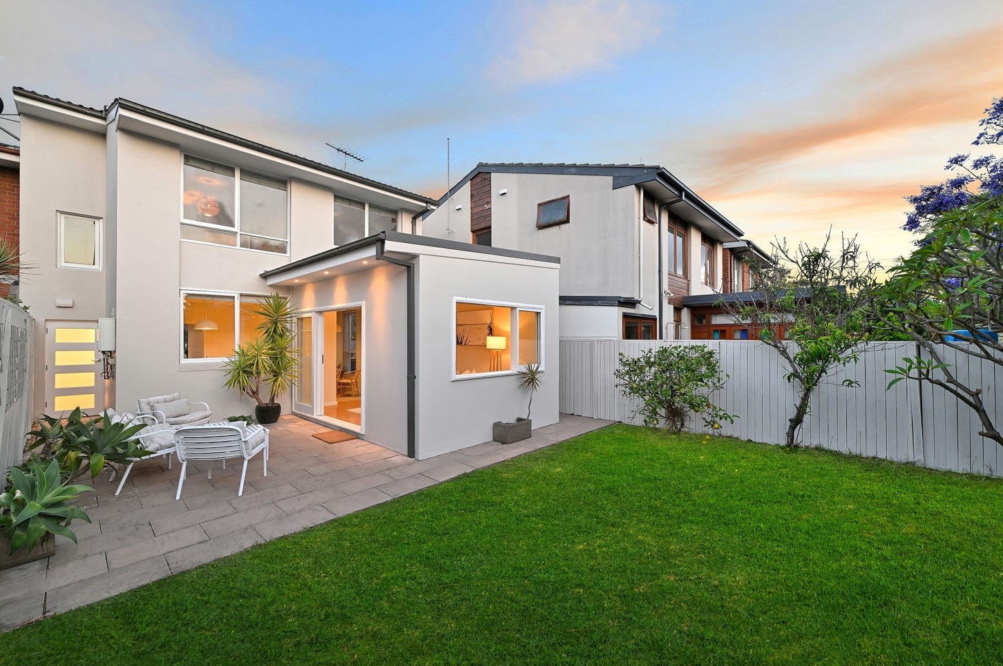 Rear of the house at dusk: stone terrace with white outdoor lounge, single-storey pavilion and level lawn
