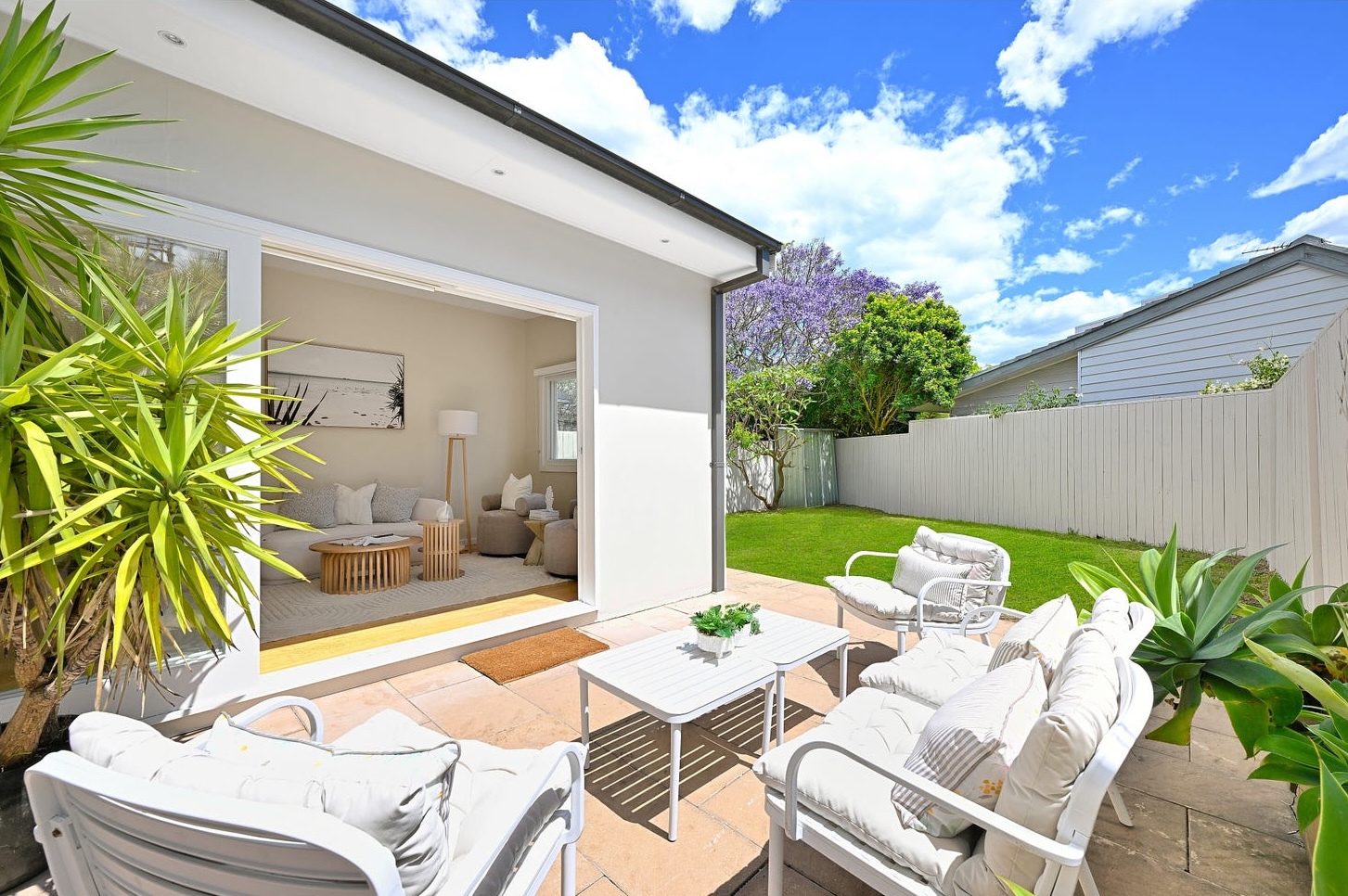 Stone terrace staged with white outdoor seating beside the rear living room, with lawn and jacaranda behind